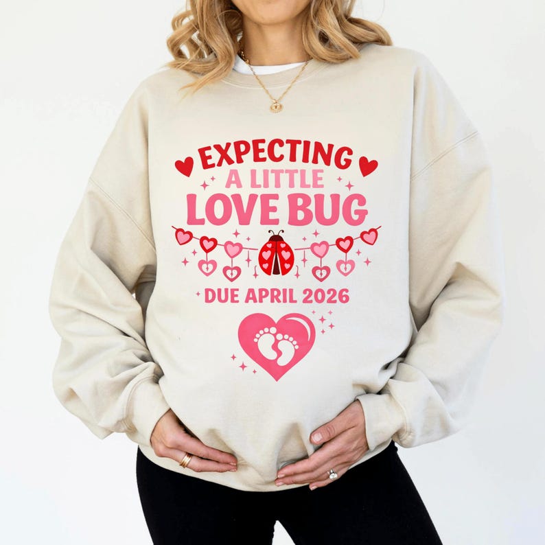 Op de afbeelding: Beige sweatshirt met de tekst "EXPECTING A LITTLE LOVE BUG" in rood en roze. Een lieveheersbeestje-afbeelding is gecentreerd onder de tekst, met een banner van harten. De woorden "DUE APRIL 2026" staan eronder. Een roze hart met voetafdrukken staat onderaan.