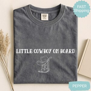 May include: A gray Comfort Colors t-shirt with the text "LITTLE COWBOY ON BOARD" in white. Below the text is a white graphic of a cowboy hat and boots. The shirt is folded and displayed on a neutral surface.