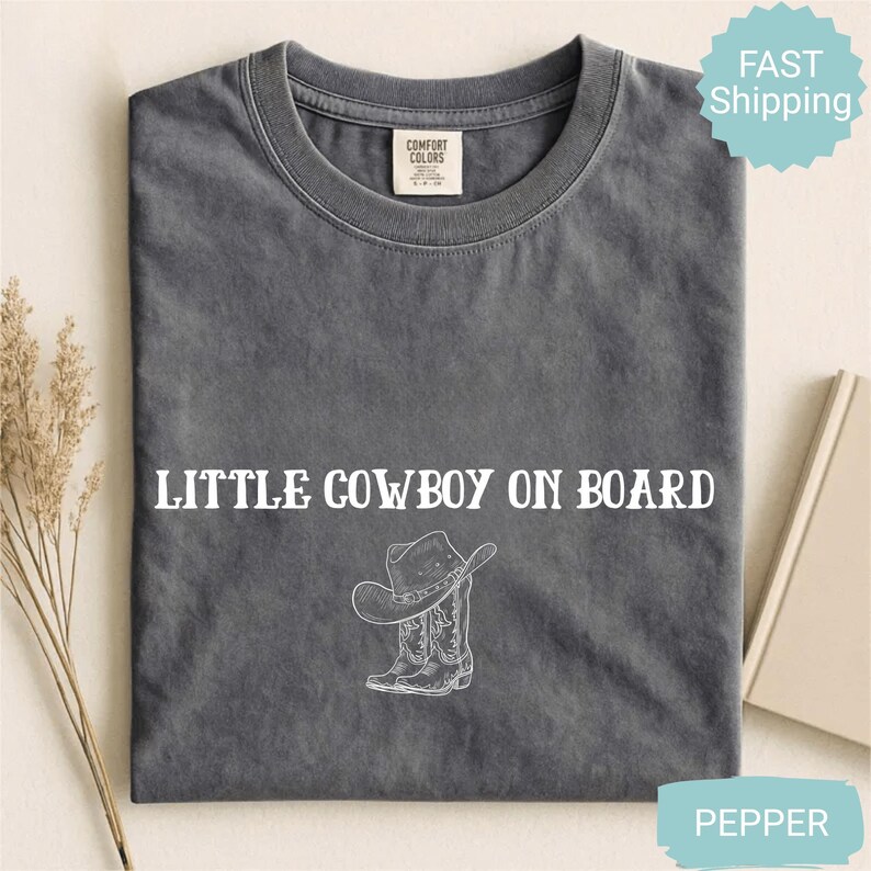 May include: A gray Comfort Colors t-shirt with the text "LITTLE COWBOY ON BOARD" in white. Below the text is a white graphic of a cowboy hat and boots. The shirt is folded and displayed on a neutral surface.