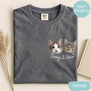 May include: A gray t-shirt featuring two cat faces with the names "Daisy & Violet" in white script. The shirt has a crew neck and a small tag near the neckline. The shirt is labeled "Pepper".
