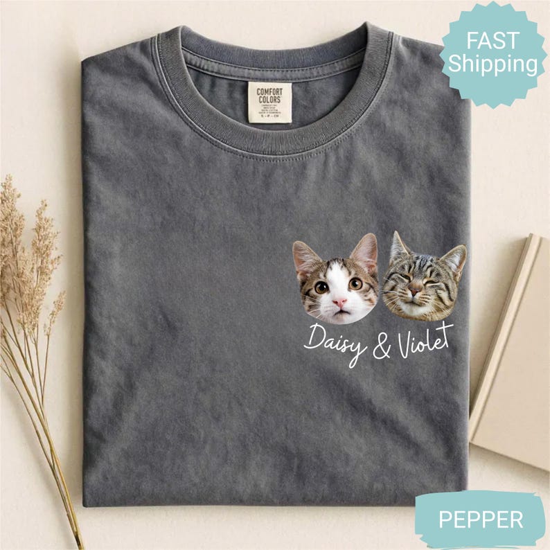 May include: A gray t-shirt featuring two cat faces with the names "Daisy & Violet" in white script. The shirt has a crew neck and a small tag near the neckline. The shirt is labeled "Pepper".