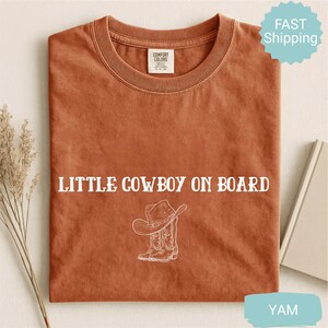 May include: A rust-colored t-shirt with the text "LITTLE COWBOY ON BOARD" in white, along with a graphic of a cowboy hat and boots. The shirt is made of Comfort Colors material.