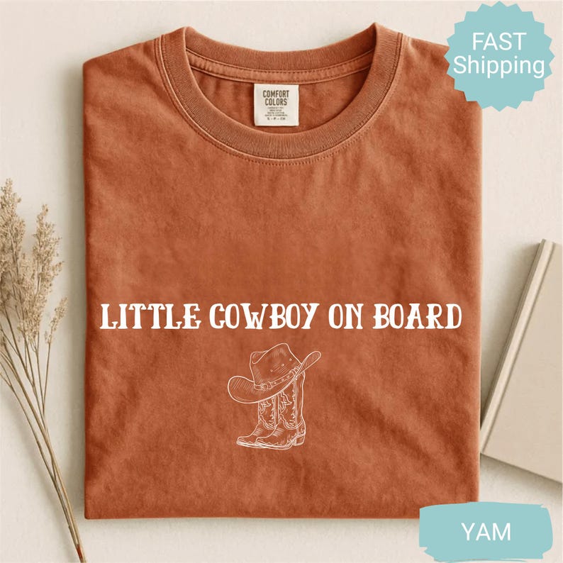 May include: A rust-colored t-shirt with the text "LITTLE COWBOY ON BOARD" in white, along with a graphic of a cowboy hat and boots. The shirt is made of Comfort Colors material.