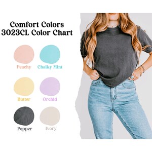 Puede incluir: Carta de colores Comfort Colors 3023CL con seis tonos pastel: Peachy, Chalky Mint, Butter, Orchid, Pepper e Ivory. Una persona lleva una camiseta gris oscuro y vaqueros azules.