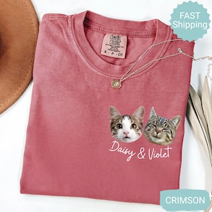 May include: Crimson-colored t-shirt featuring two cat faces with the names "Daisy & Violet" in white script. The shirt has a crew neck and a small tag at the neckline. A silver necklace with a pendant is draped across the shirt.