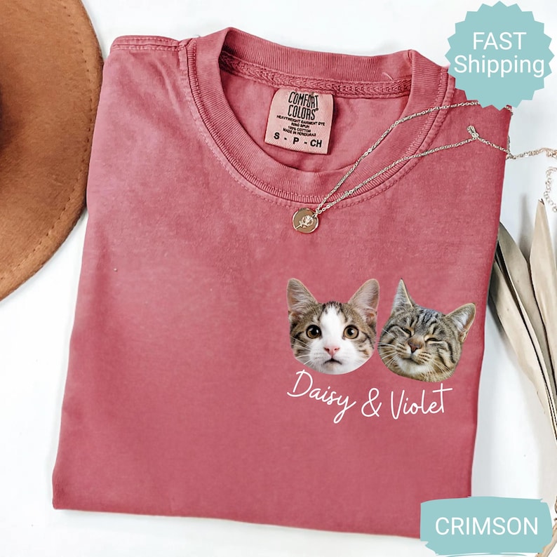 May include: Crimson-colored t-shirt featuring two cat faces with the names "Daisy & Violet" in white script. The shirt has a crew neck and a small tag at the neckline. A silver necklace with a pendant is draped across the shirt.
