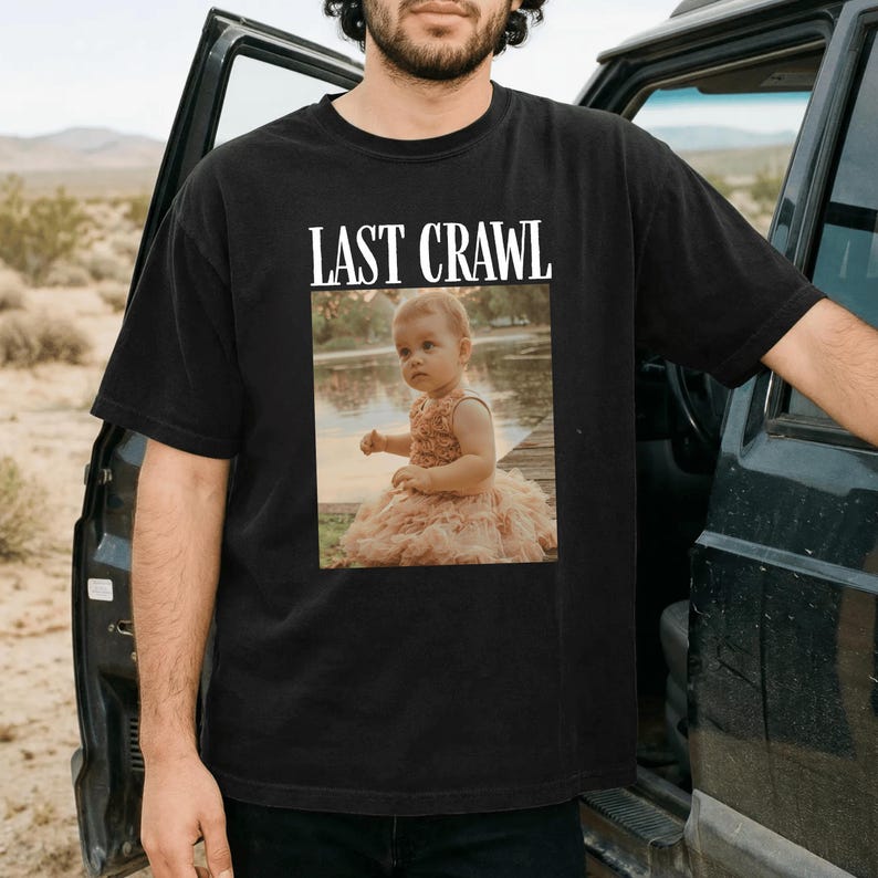 May include: Black t-shirt featuring the text "LAST CRAWL" above a photograph of a baby in a pink dress. The baby is seated on a wooden dock near water. The t-shirt is being worn by a person.
