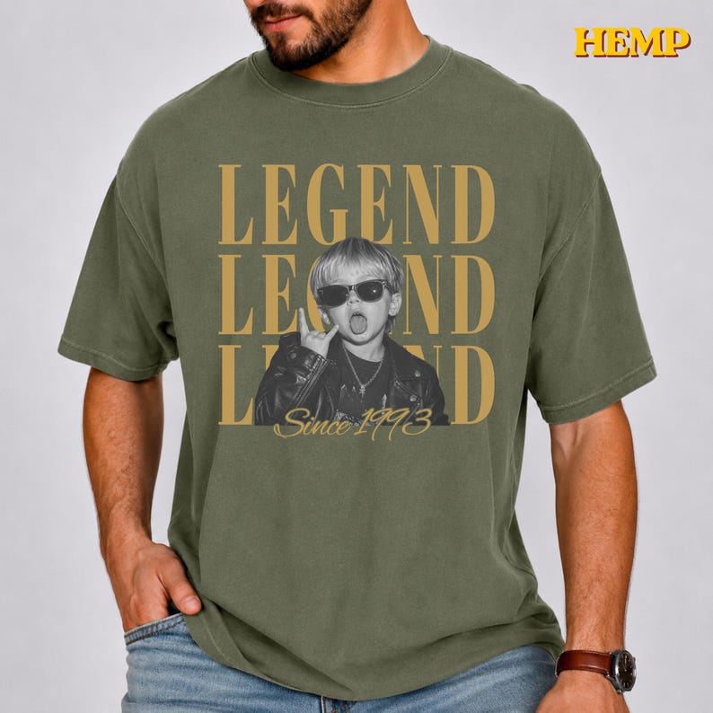 Puede incluir: Una camiseta verde oliva con el texto HEMP en la esquina superior derecha. El gr&aacute;fico central muestra la palabra LEGEND en letras doradas grandes, una foto en blanco y negro de un ni&ntilde;o con gafas de sol y chaqueta de cuero, y Since 1993.