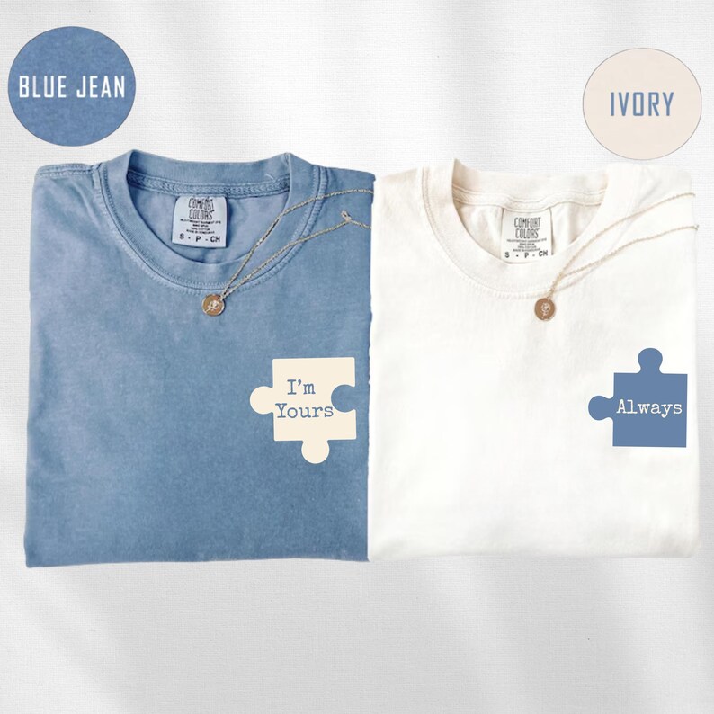 May include: Two t-shirts, one blue and one ivory, with matching puzzle piece graphics. The blue shirt says "I'm Yours" and the ivory shirt says "Always". Each shirt has a necklace. The blue shirt is labeled "Blue Jean" and the ivory shirt is labeled "Ivory".