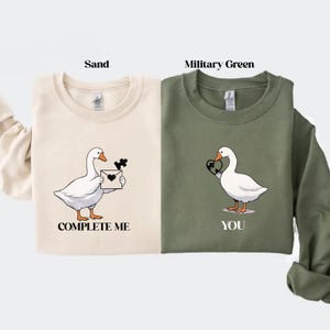 May include: Sand-colored and military green sweatshirts with goose graphics. The sand sweatshirt has a goose holding a puzzle piece and a letter, with the text "COMPLETE ME". The green sweatshirt has a goose holding a heart, with the text "YOU".