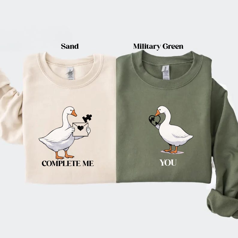 May include: Sand-colored and military green sweatshirts with goose graphics. The sand sweatshirt has a goose holding a puzzle piece and a letter, with the text "COMPLETE ME". The green sweatshirt has a goose holding a heart, with the text "YOU".
