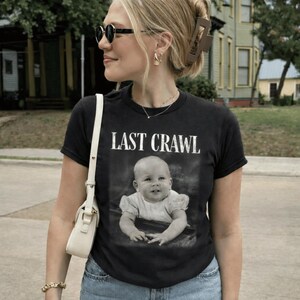 May include: Black t-shirt with the text "LAST CRAWL" above a black and white image of a baby. The person is wearing sunglasses, a gold necklace, and a white purse. The background includes a green house and a street.