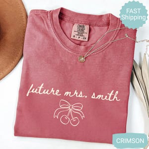 May include: Crimson-colored t-shirt with the text "future mrs. smith" and a cherry bow graphic. The shirt has a crew neck and short sleeves. A gold necklace with a pendant is draped across the shirt.