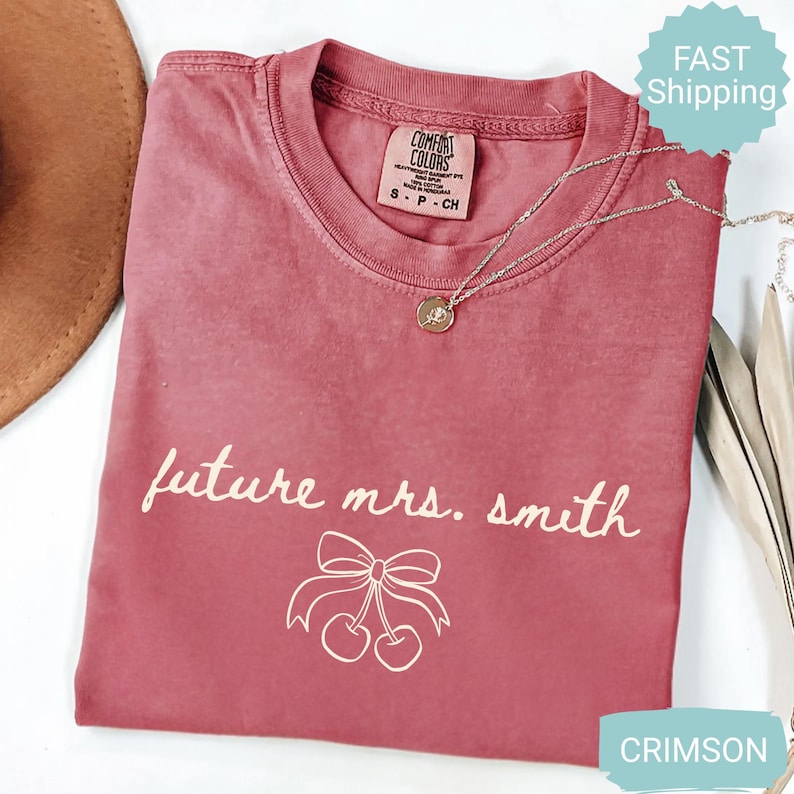 May include: Crimson-colored t-shirt with the text "future mrs. smith" and a cherry bow graphic. The shirt has a crew neck and short sleeves. A gold necklace with a pendant is draped across the shirt.