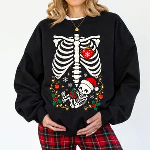 Puede incluir: Una sudadera negra de manga larga con un gráfico blanco de una caja torácica humana y un adorno de corazón rojo. Debajo, un pequeño esqueleto con gorro de Papá Noel sostiene un regalo verde, rodeado de copos de nieve y luces navideñas.