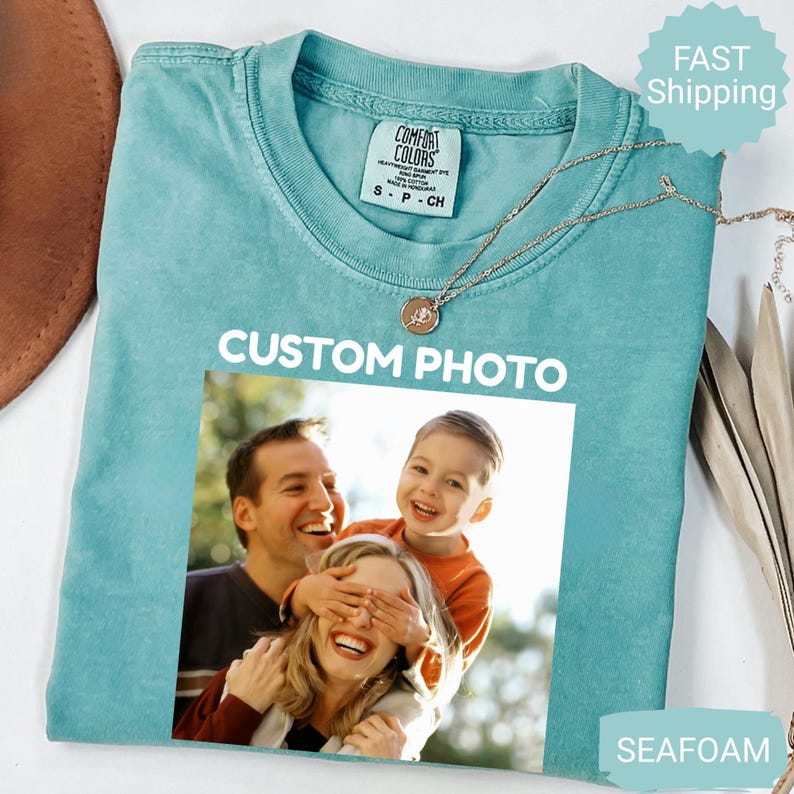 May include: Seafoam green Comfort Colors t-shirt with a photo print. The shirt features the text "CUSTOM PHOTO" in white. A gold necklace with a pendant is on the shirt. The photo shows a family of three. The shirt has a tag that says "Comfort Colors".
