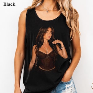 Puede incluir: Una camiseta sin mangas negra con un estampado gr&aacute;fico de una persona con cabello largo casta&ntilde;o que lleva un top estilo cors&eacute; marr&oacute;n oscuro y un collar. La camiseta tiene un cuello redondo y un corte relajado con un dise&ntilde;o detallado.