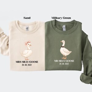Peut inclure: Deux sweatshirts, un beige et un vert militaire, chacun avec une illustration d'oie. Le sweat-shirt beige a un n&oelig;ud rose sur l'oie et porte l'inscription "MRS SILLY GOOSE 20.08.2022". Le sweat-shirt vert porte l'inscription "MR SILLY GOOSE 20.08.2022".