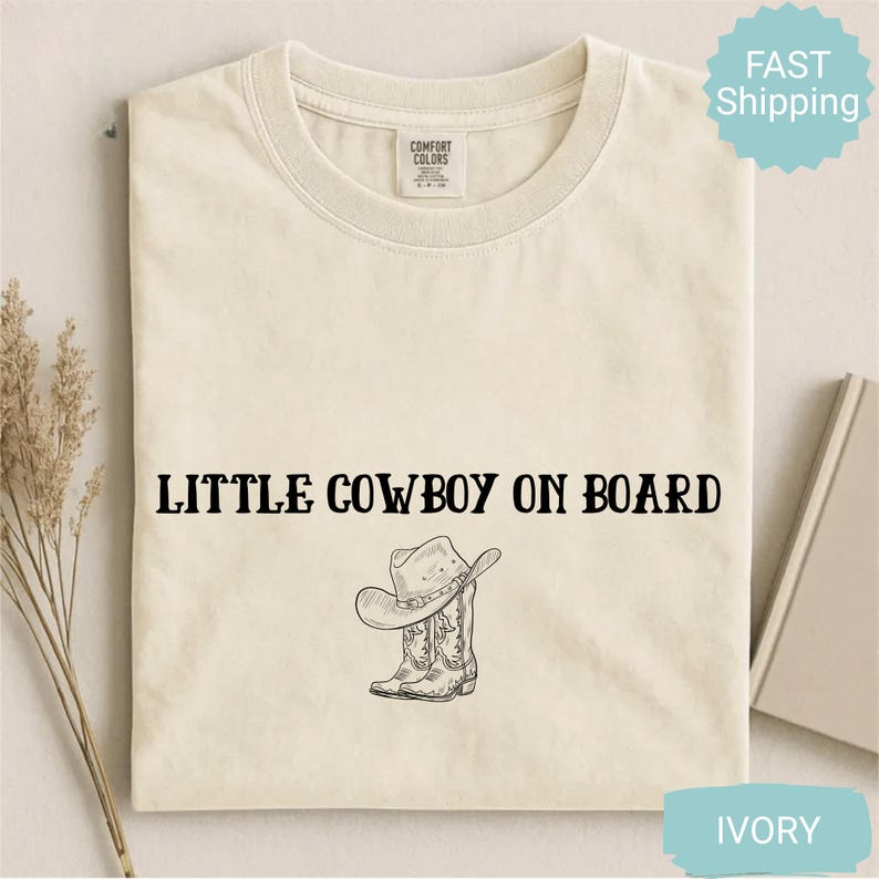 May include: Ivory-colored t-shirt with the text "LITTLE COWBOY ON BOARD" in black. Below the text is a graphic of a cowboy hat and boots. The shirt has a crew neck and a tag on the upper back.