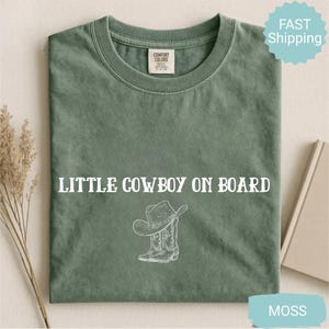 May include: A moss green t-shirt with the text "LITTLE COWBOY ON BOARD" in white. Below the text is a white illustration of a cowboy hat and boots. The shirt has a crew neck and short sleeves.