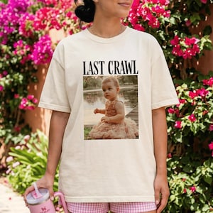 May include: A cream-coloured t-shirt with the words "LAST CRAWL" printed above a photograph of a baby in a dress. The baby is near water. The person wearing the shirt is holding a pink tumbler.