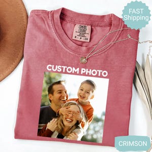May include: Crimson-colored t-shirt with a crew neck and a photo print. The shirt features the text "CUSTOM PHOTO" above a family photo. A silver necklace with a pendant is draped on the shirt.