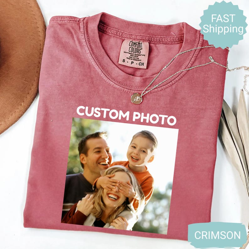 May include: Crimson-colored t-shirt with a crew neck and a photo print. The shirt features the text "CUSTOM PHOTO" above a family photo. A silver necklace with a pendant is draped on the shirt.