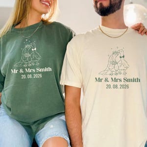 May include: Two matching shirts, one in olive green and the other in cream, feature a line drawing of two doves with floral accents. The text "Mr & Mrs Smith 20.08.2026" is printed below the graphic, ideal for a wedding or anniversary.