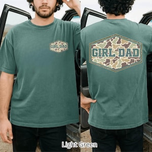 May include: Light green t-shirt with "GIRL DAD" text in a camouflage design. The shirt has a crew neck and short sleeves. The text is in a hexagonal shape on the front and back of the shirt.