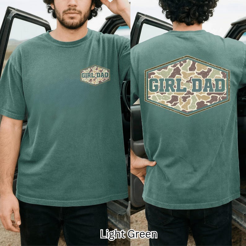 May include: Light green t-shirt with "GIRL DAD" text in a camouflage design. The shirt has a crew neck and short sleeves. The text is in a hexagonal shape on the front and back of the shirt.