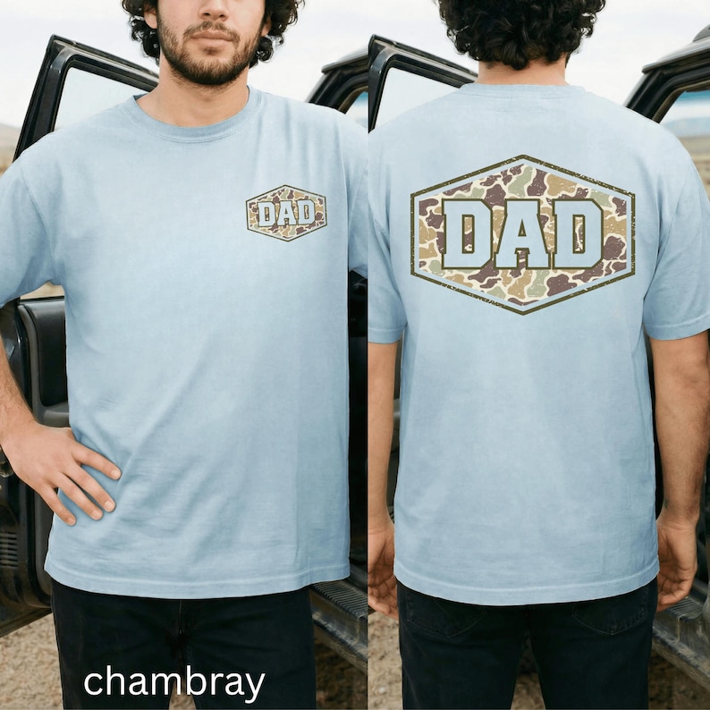 May include: Light blue chambray t-shirt with a front and back graphic. The front graphic is a brown and green camouflage design with the word "DAD" in light blue. The back graphic is a larger version of the same design.