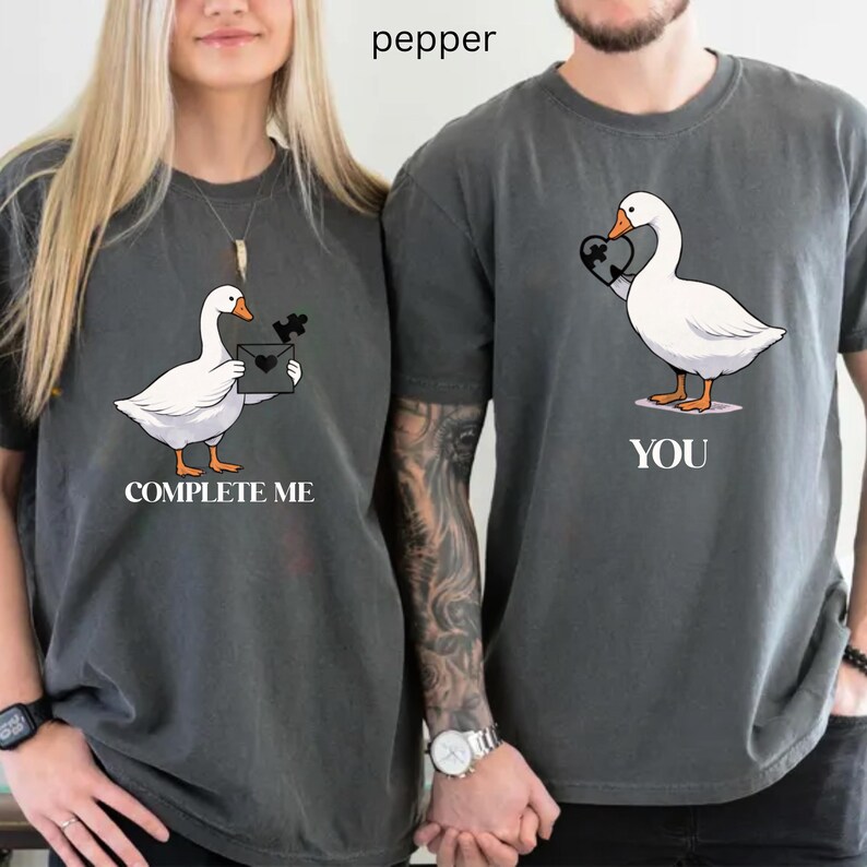 May include: Two matching gray t-shirts with goose graphics. One shirt says "COMPLETE ME" and features a goose holding a puzzle piece. The other shirt says "YOU" and has a goose holding a heart.