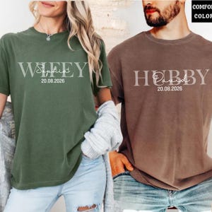 May include: Two short-sleeved t-shirts, one olive green and one brown. The olive green shirt says "Wifey Sophia 20.08.2026" in a light gray font. The brown shirt says "Hubby David 20.08.2026" in a light gray font.