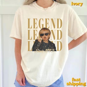 Puede incluir: Una camiseta de manga corta color marfil con un estampado gr&aacute;fico de un ni&ntilde;o con chaqueta de cuero negra y gafas de sol, haciendo un gesto con la mano. El texto "LEGEND" se repite en letras doradas grandes, con "Since 1993" en cursiva.