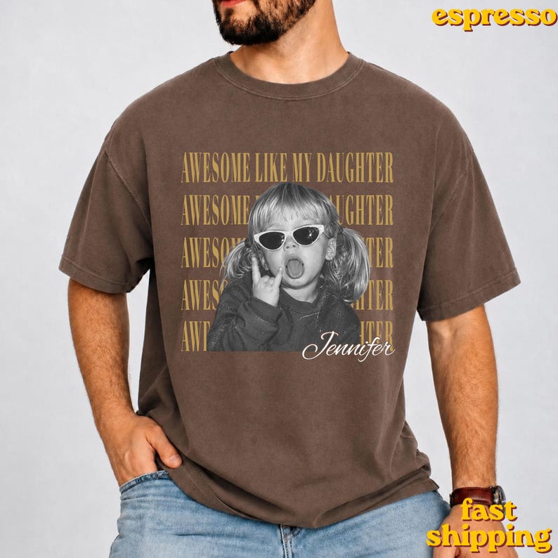 May include: A brown short-sleeve t-shirt featuring a black and white photograph of a child wearing sunglasses and making a hand gesture. The text AWESOME LIKE MY DAUGHTER is repeated in gold, and the name Jennifer is written in white script.