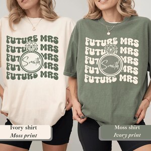 K&ouml;nnte beinhalten: Zwei T-Shirts, eines in Elfenbein und eines in Moosgr&uuml;n, mit dem sich wiederholenden Text "FUTURE MRS". Eine Diamantring-Grafik umgibt den Namen "Smith". Das elfenbeinfarbene Shirt hat einen moosgr&uuml;nen Aufdruck, und das moosgr&uuml;ne Shirt hat einen elfenbeinfarbenen Aufdruck.