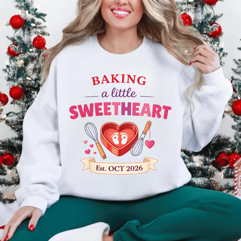 Op de afbeelding: Witte sweatshirt met de tekst "BAKING a little SWEETHEART" in rood en roze. Het ontwerp bevat een hartvormige bakvorm met voetafdrukken, gardes en de tekst "Est. OCT 2026" op een banner.