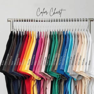 May include: A colour chart showing a range of t-shirts in various colours, including black, navy, red, yellow, pink, green, blue, and white. The shirts are hanging on a metal rack with the words "Color Chart" in cursive.