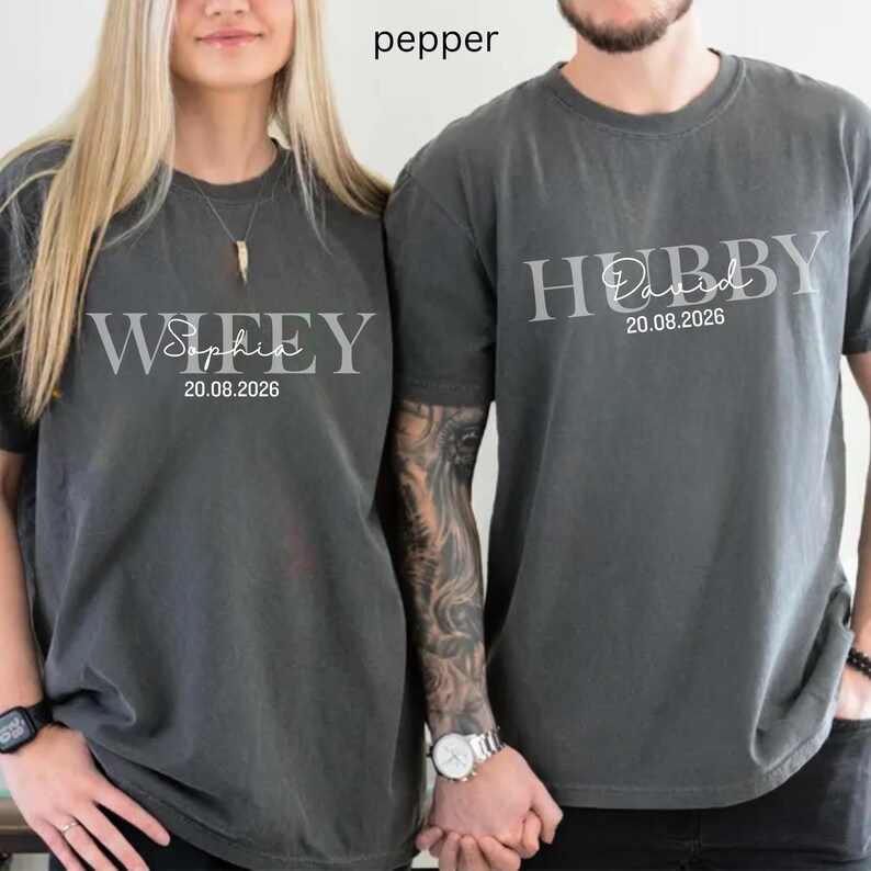 May include: Two matching pepper-colored t-shirts. One shirt says "Wifey Sophia 20.08.2026" in a light gray font. The other shirt says "Hubby Daniel 20.08.2026" in a light gray font. The shirts are short-sleeved and have a crew neck.