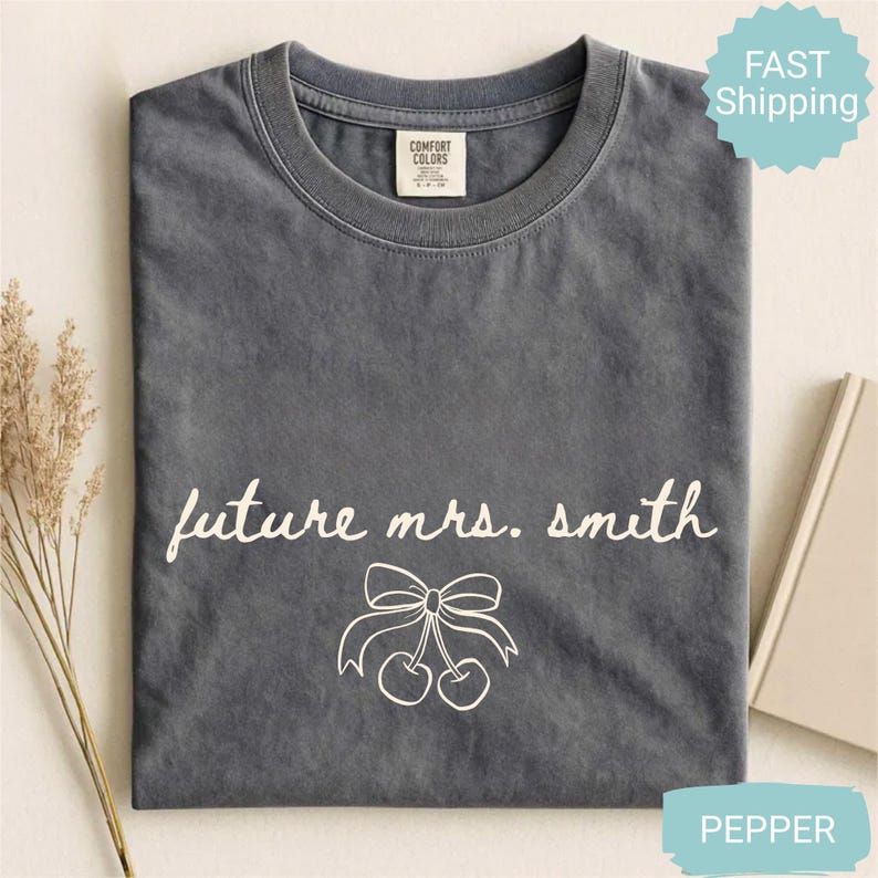 May include: A gray Comfort Colors t-shirt with the text "future mrs. smith" in white script, along with a bow and cherry graphic. The shirt is folded and has a tag in the upper center. The word "pepper" is visible in the bottom right corner.