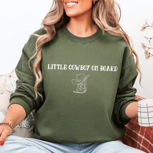 May include: Olive green sweatshirt with the text "LITTLE COWBOY ON BOARD" in white, along with a white graphic of a cowboy hat and boots. The sweatshirt is a crew neck style.