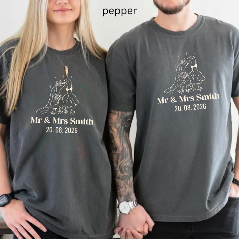May include: Two pepper-colored t-shirts with a white graphic of two birds and the text "Mr & Mrs Smith 20.08.2026". The shirts are short-sleeved and have a relaxed fit. The shirts are worn by two people.