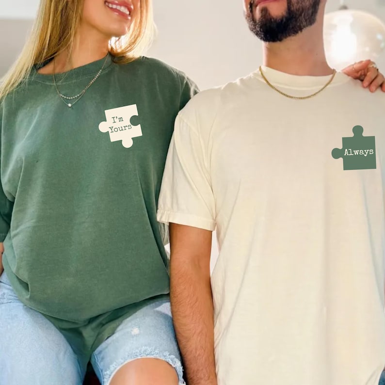 May include: Two matching t-shirts. One is a dark green sweatshirt with the text "I'm Yours" inside a white puzzle piece. The other is a cream-colored t-shirt with the text "Always" inside a green puzzle piece.