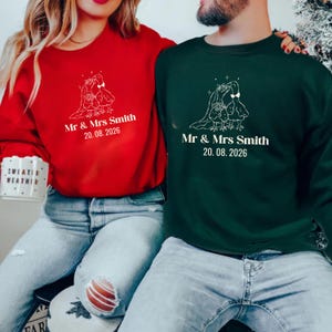 May include: Red and green crewneck sweatshirts with matching white graphic of two birds with a wedding veil and the text "Mr & Mrs Smith 20.08.2026". The sweatshirts are worn by a couple, suggesting a wedding or anniversary theme.