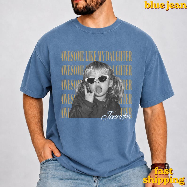 May include: A blue jean coloured t-shirt featuring a graphic of a child wearing sunglasses and making a hand gesture. The text reads AWESOME LIKE MY DAUGHTER repeated in gold, with the name Jennifer written in white script below the child.