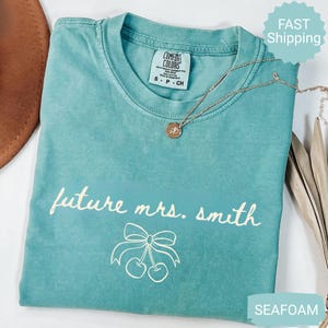 May include: Seafoam green t-shirt with the text "future mrs. smith" and a bow with cherries graphic. A gold necklace with a pendant is on the shirt. The shirt has a Comfort Colors tag.