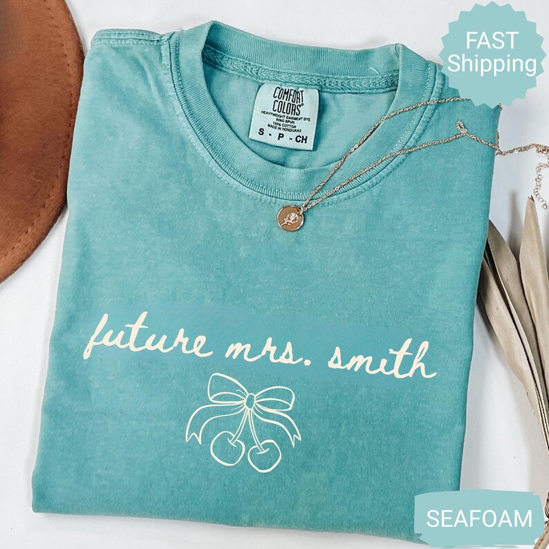 May include: Seafoam green t-shirt with the text "future mrs. smith" and a bow with cherries graphic. A gold necklace with a pendant is on the shirt. The shirt has a Comfort Colors tag.