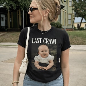 May include: Black t-shirt featuring the text "LAST CRAWL" and a vintage baby photo. The shirt is worn by a person wearing sunglasses and a white shoulder bag.