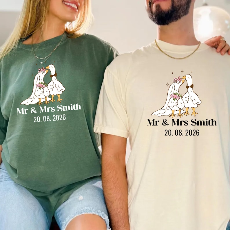 May include: Two t-shirts, one green and one cream, featuring a cartoon illustration of two geese with floral accents. The text "Mr & Mrs Smith" and the date "20.08.2026" are printed below the illustration. The shirts are likely for a wedding or anniversary.