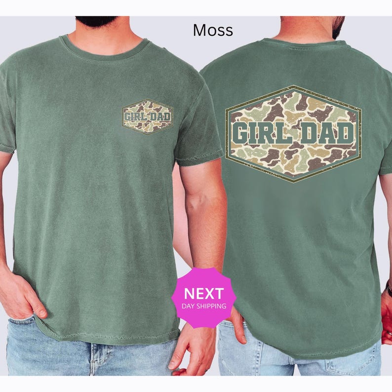 May include: Moss green t-shirt with "GIRL DAD" text and camouflage design on the front and back. The shirt has a crew neck and short sleeves. The text is in a distressed font, and the camouflage pattern includes shades of green, brown, and tan.